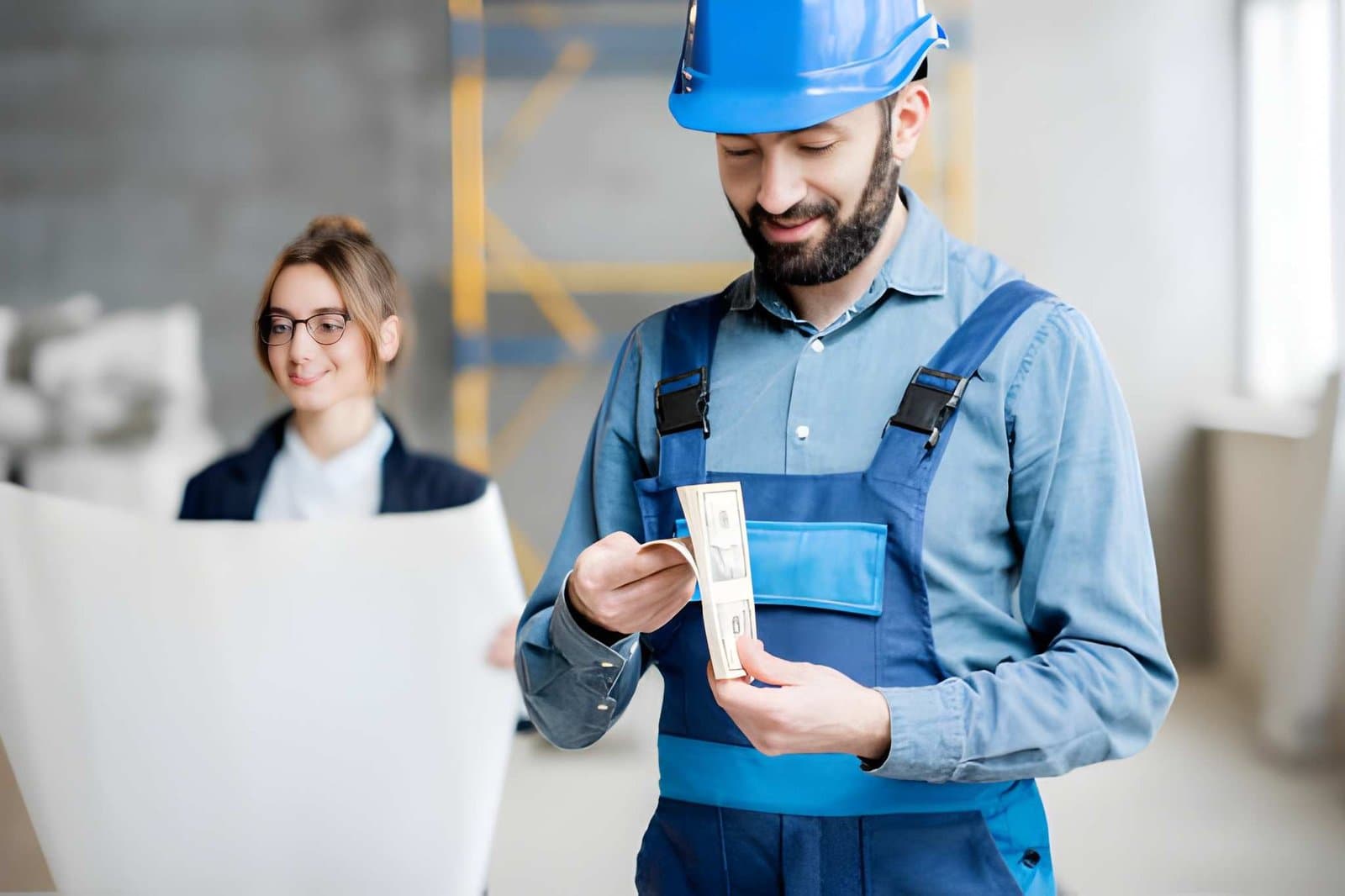 Construction manager in a hard hat counting banknotes on site while a colleague reviews plans