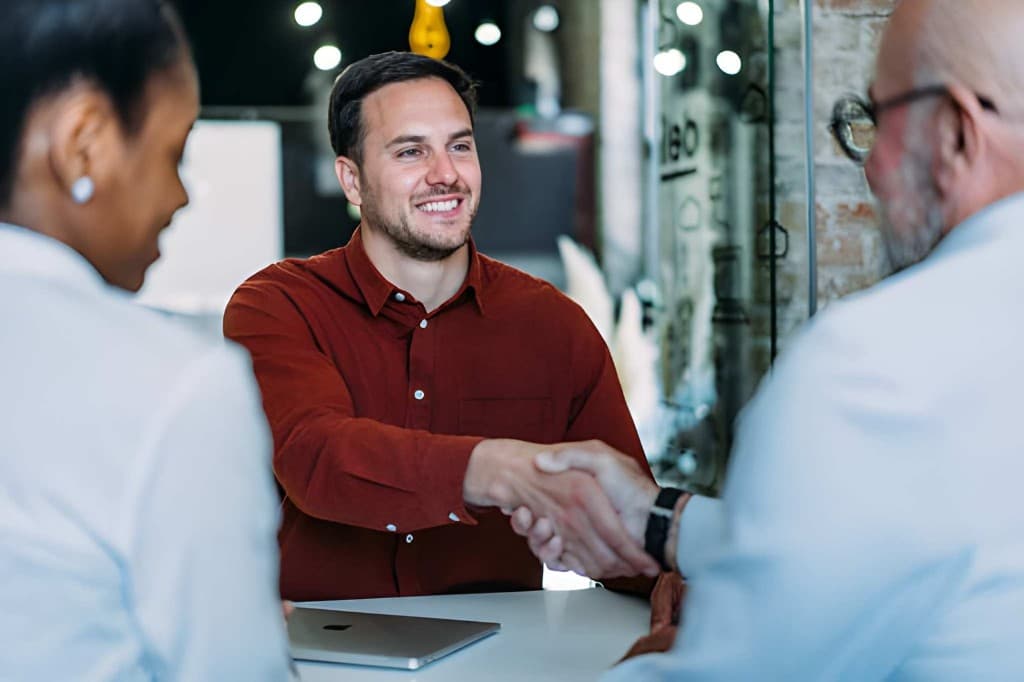 Smiling candidate in a rust-coloured shirt shaking hands across a table during a job interview