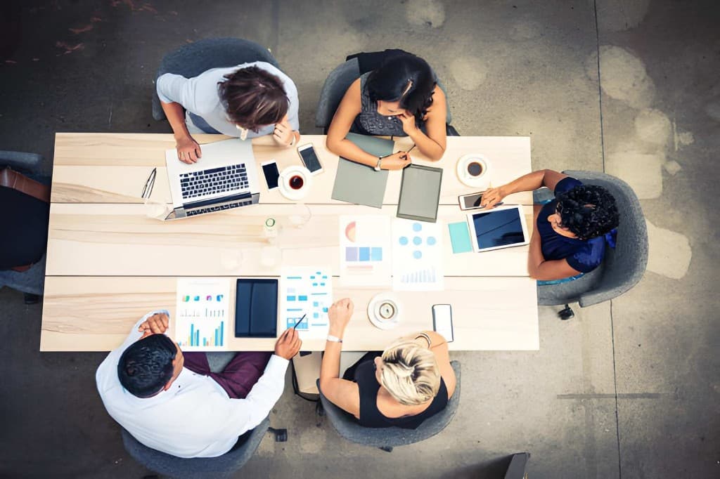 Top-down view of five professionals collaborating around a table with laptops, tablets and data documents